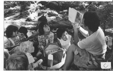 Children in bush with teacher (right) reading story book to some of them - Resource cover image